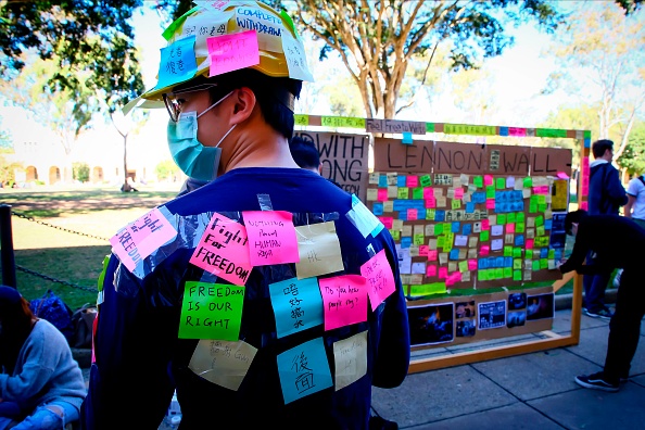 -Cette photo prise le 9 août 2019 montre un défenseur des manifestations pro-démocratiques de Hong Kong, couvert de notes autocollantes, debout devant un "mur de Lennon" improvisé à l'Université du Queensland à Brisbane en Australie. Les tensions à Hong Kong ont eu des répercussions sur les campus universitaires australiens, provoquant des affrontements entre étudiants pro-démocrates et pro-pékinois, allant du harcèlement aux menaces de mort. Photo de Patrick HAMILTON / AFP / Getty Images.