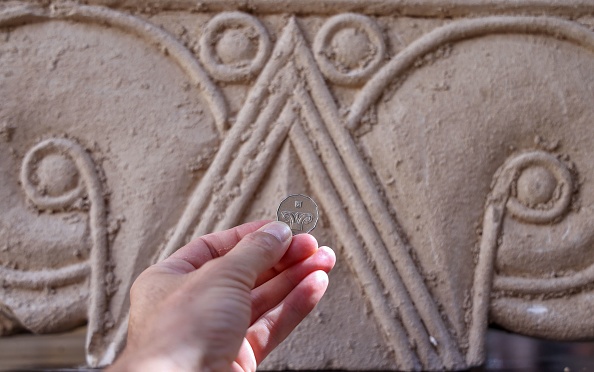 -Une pièce de monnaie israélienne de cinq shekels est tenue devant un chapiteau de colonne affichant les mêmes sculptures décoratives. Photo par EMMANUEL DUNAND / AFP via Getty Images.