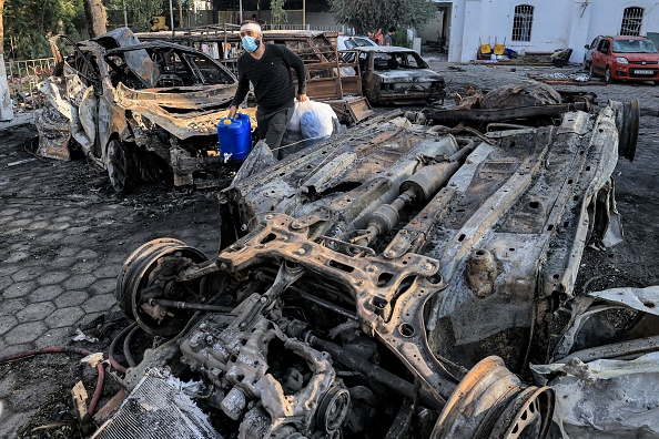 Un homme marche avec des objets récupérés devant des véhicules détruits sur le site de l'hôpital Ahli Arab dans la ville de Gaza, le 18 octobre 2023.   (MAHMUD HAMS/AFP via Getty Images)