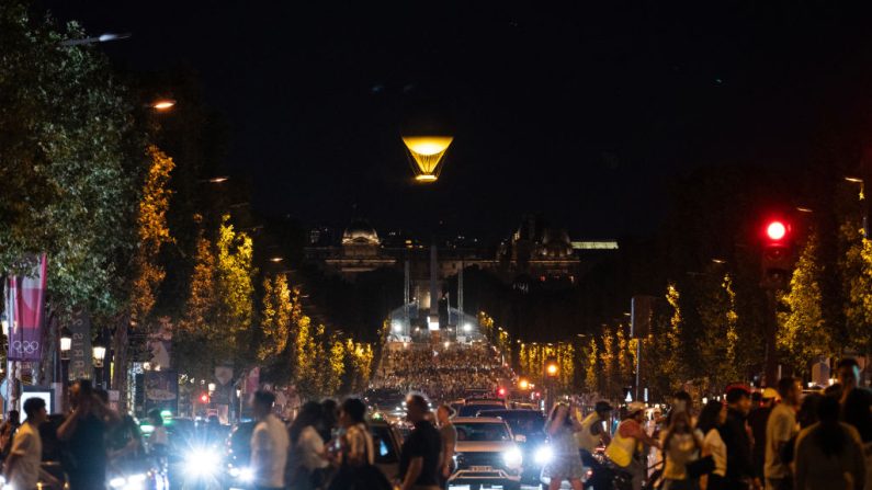 Dans cette vue de l'avenue des Champs-Élysées, des piétons se rassemblent alors que la vasque des Jeux olympiques et paralympiques de Paris 2024, attachée à un ballon, est soulevée dans les airs au-dessus du jardin des Tuileries à Paris, le 8 août 2024, pendant les JO de Paris 2024. (Crédit photo STEFANO RELLANDINI/AFP via Getty Images)