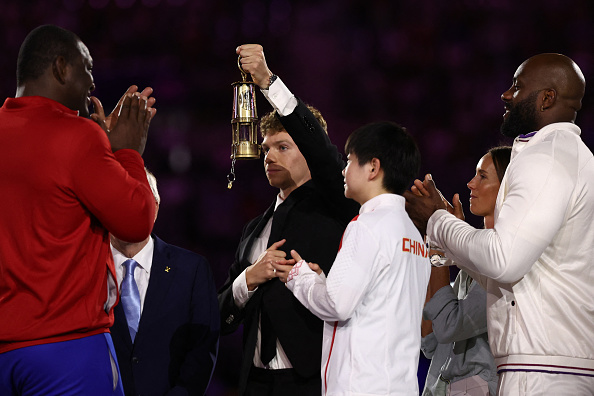 Le nageur Léon Marchand arrive avec la flamme olympique et la montre aux athlètes, dont le judoka français Teddy Riner (à dr.), lors de la cérémonie de clôture des Jeux olympiques de Paris 2024 au Stade de France, à Saint-Denis, le 11 août 2024. (Photo FRANCK FIFE/AFP via Getty Images)