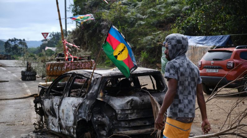 
Un manifestant indépendantiste kanak passe devant un drapeau kanak et du Front de libération nationale socialiste (FLNKS) placé sur une voiture endommagée lors d'un barrage routier à Houailou, sur la côte est du territoire français du Pacifique de la Nouvelle-Calédonie, le 1er juillet 2024. (THEO ROUBY/AFP via Getty Images)